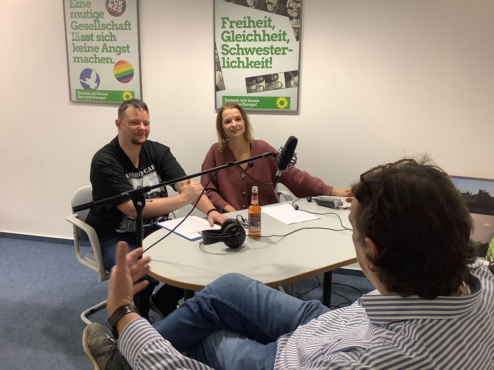 Blick in die Podcast-Aufnahme York und Jasmin in Gespräch mit Hartmut. Im HIntergrud zwei Plakate "Eine mutige GEsellschaft lässt sich keine Angst machen" und "Freiheit - Gleichheit - Schwesterlichkeit