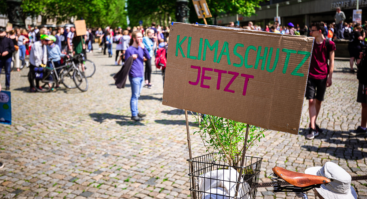 Ein Schild mit der Aufschrift „Klimaschutz jetzt“ mit demonstrierenden Menschen im Hintergrund.