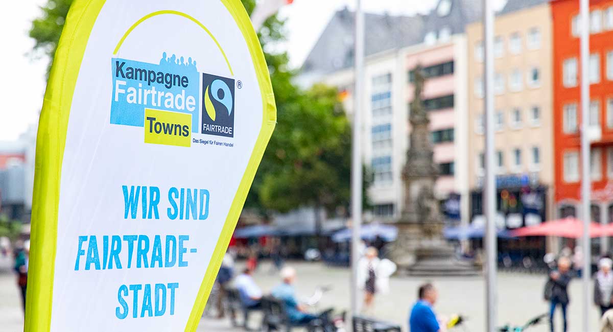 Eine Beachflag im Fairtrade-Branding vor einer unscharfen Stadtkulisse.