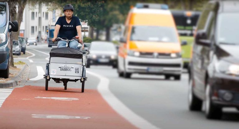 Ein Lastenradfahrer auf einem roten Radweg wird von Autos überholt.