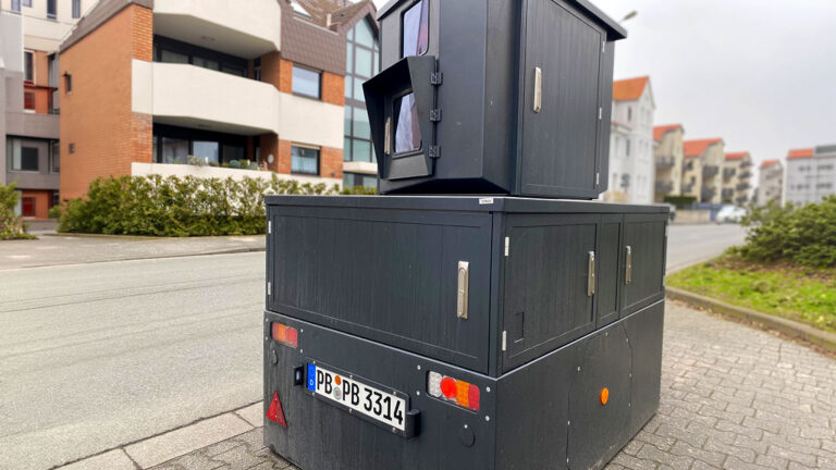 Schwarzer Radaranhänger am Straßenrand vor einer Häuserzeile.
