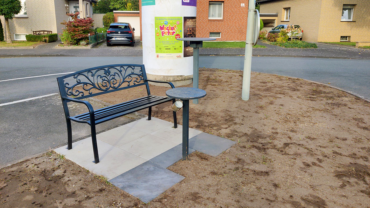 Schwarze Sitzbank aus Metall mit Tischchen auf einer gepflasterten Fläche.