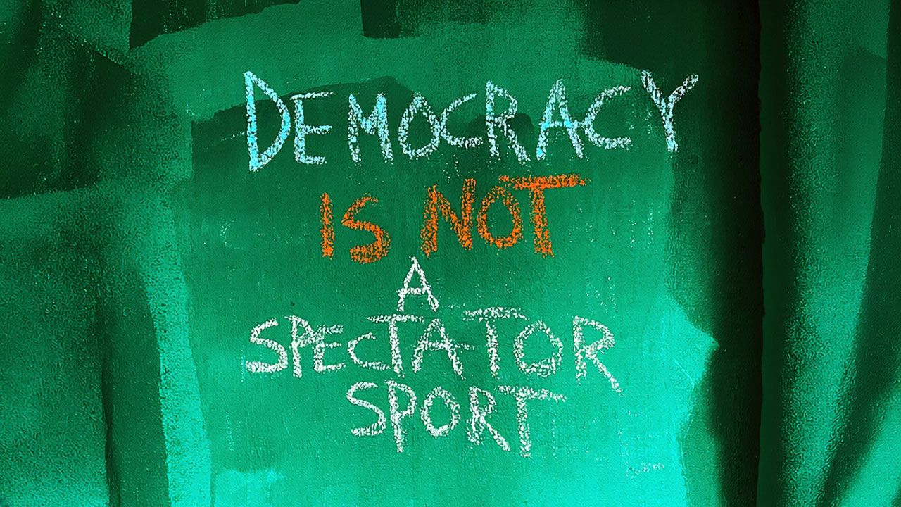 Auf einer grünen Wand steht der Schriftzug DEMOCRACY IS NOT A SPECTATOR SPORT.